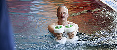 Ein Mann steht mit zwei Hanteln in der Hand in einem Schwimmbecken.